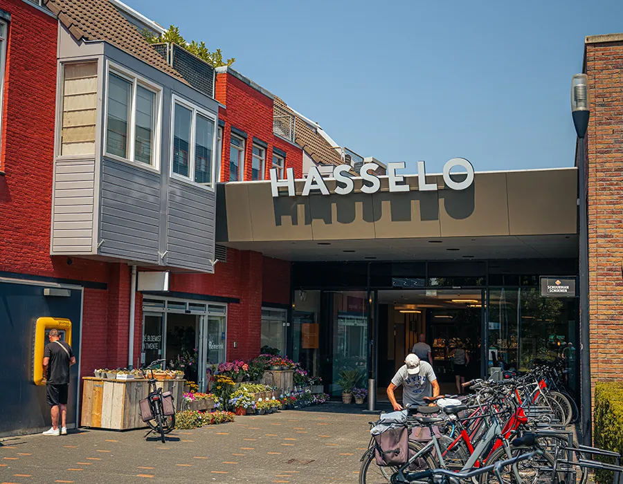 Ingang van Winkelcentrum Hasselo met het grote Hasselo-logo, een bloemenmarkt aan de zijkant en fietsen geparkeerd bij de entree