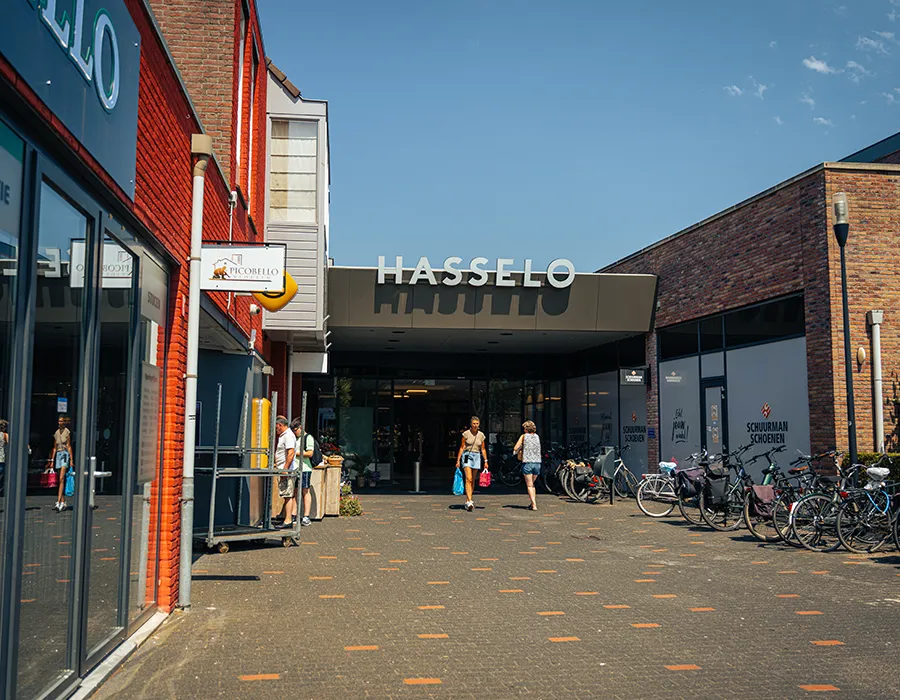 Ingang van Winkelcentrum Hasselo met het Hasselo-logo, Schuurman Schoenen rechts en bezoekers die het winkelcentrum in- en uitlopen