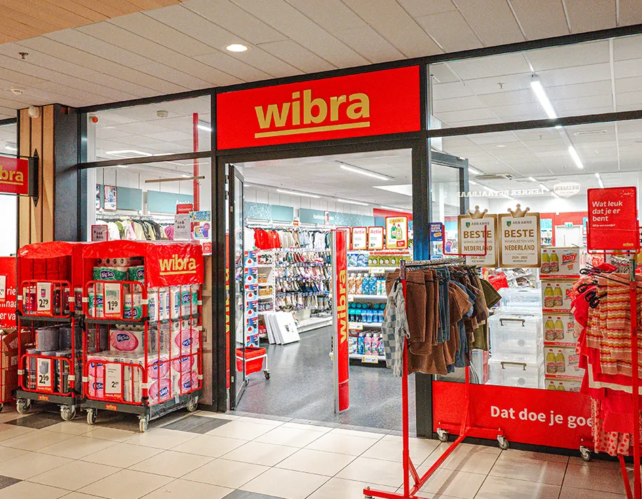 Ingang van Wibra in Winkelcentrum Hasselo met een opvallende rode gevel, winkeltrolley met aanbiedingen en de tekst 'Dat doe je goed'