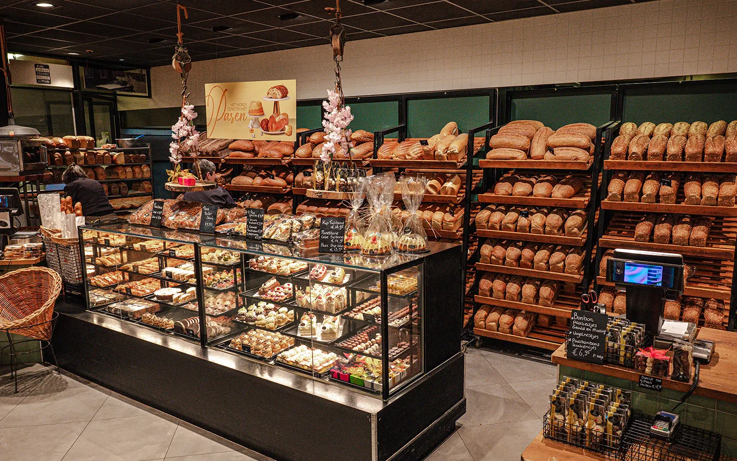 Breed interieuroverzicht van Wouda De Echte Bakker in Winkelcentrum Hasselo met een gevulde gebaksvitrine, volle broodschappen en Pasen decoratie
