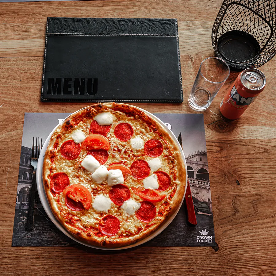 Gebakken pizza met salami, tomaat en mozzarella op een houten tafel bij Elat Grillroom en Pizzeria in Winkelcentrum Hasselo, geserveerd met een drankje en menu