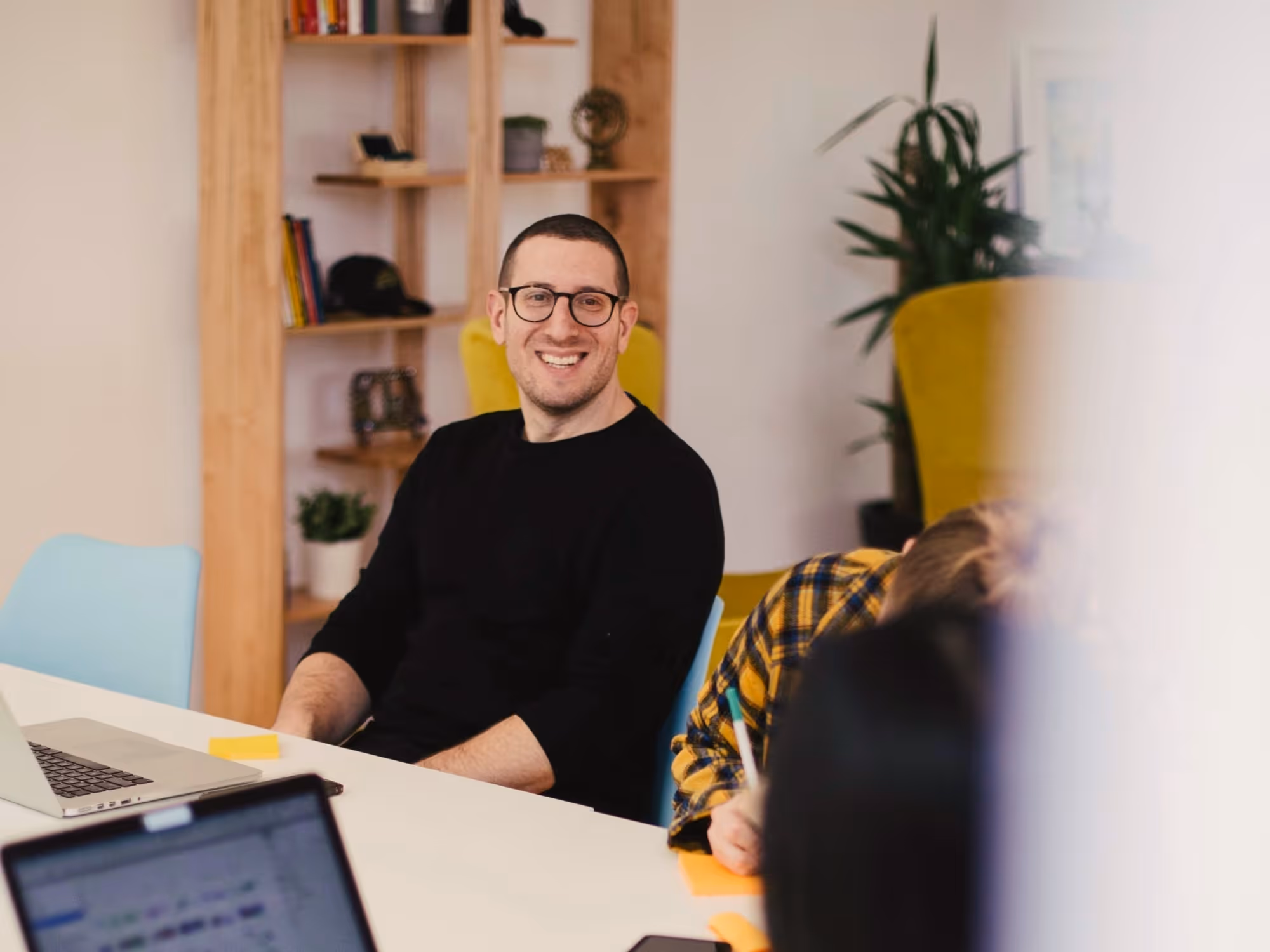 Man in glasses and black shirt smiling while sitting at a table with a laptop in a modern office setting.