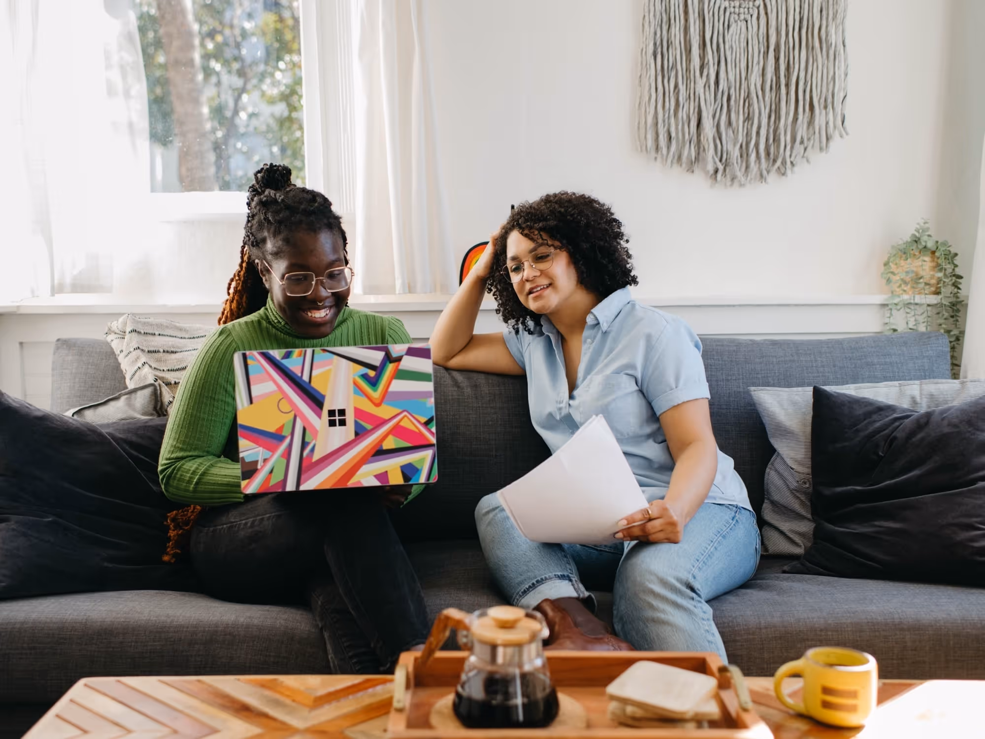 Two women sitting on a gray sofa, one with glasses and a green sweater using a colorful laptop, the other holding papers and wearing a light blue shirt and jeans.