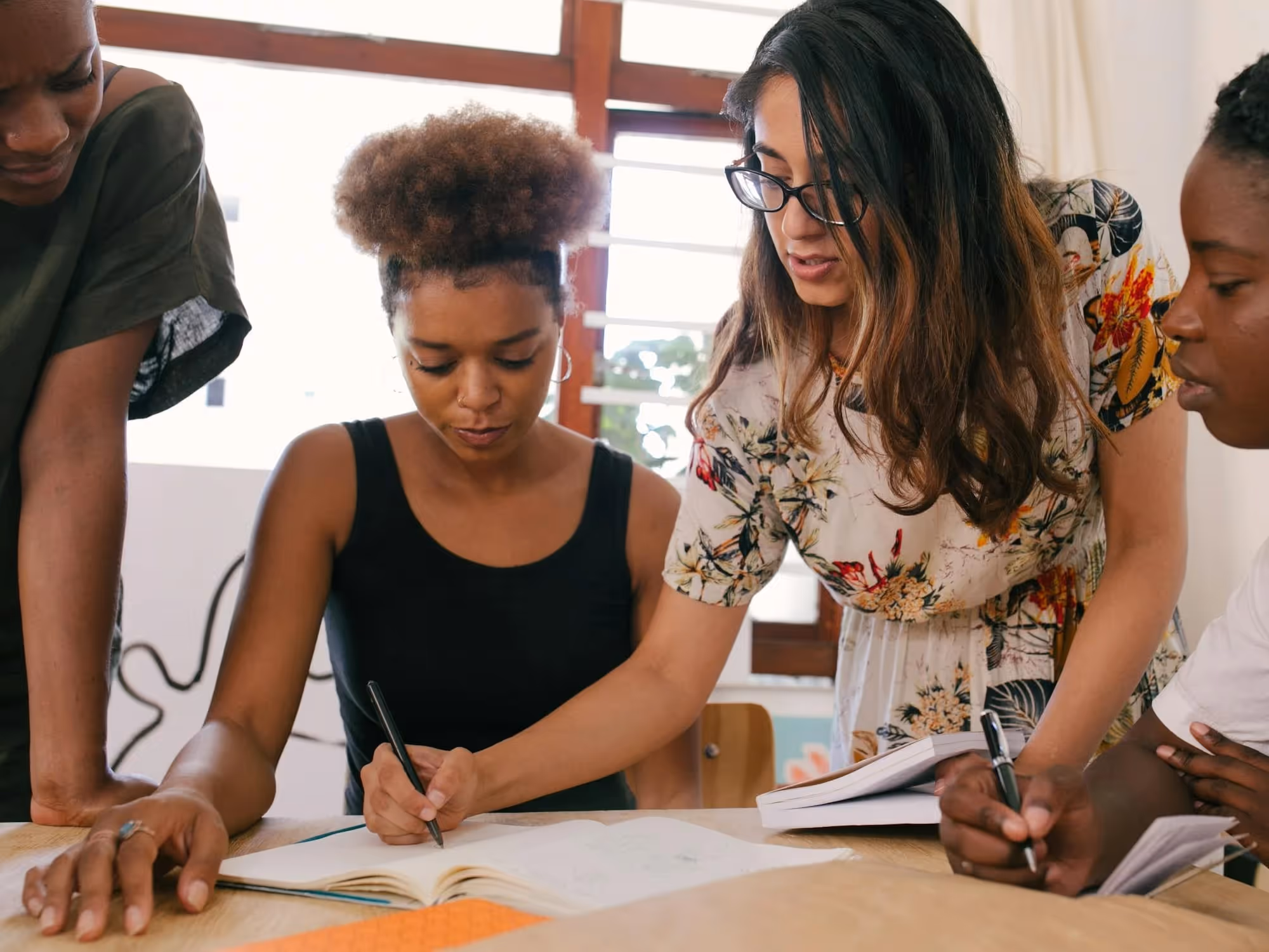 Four young women collaborating and writing in notebooks around a wooden table in a bright room.