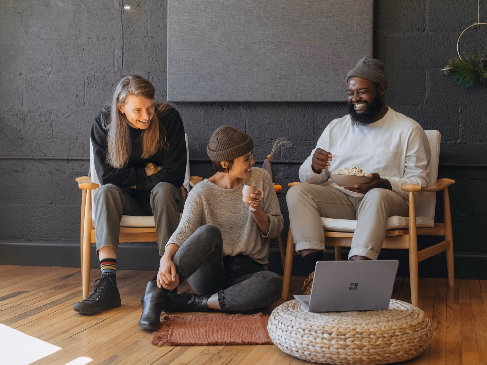 Three friends laughing and sitting around a laptop on a wicker ottoman in a cozy room with wooden floors.