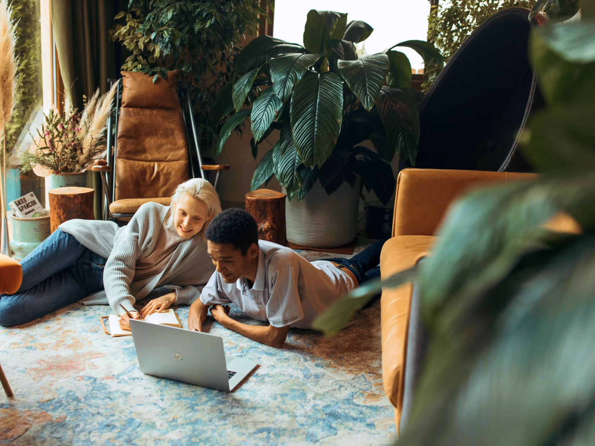 Two people lying on a colorful rug in a cozy room looking at a laptop, with a large green plant and comfortable chairs nearby.