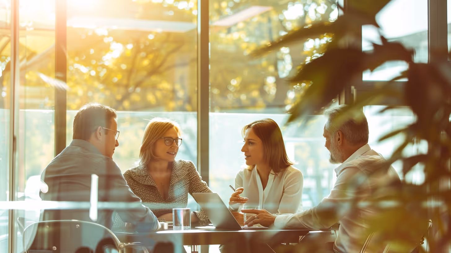 Four colleagues having a meeting around a table with a laptop and glasses of water in a sunlit office with large windows.
