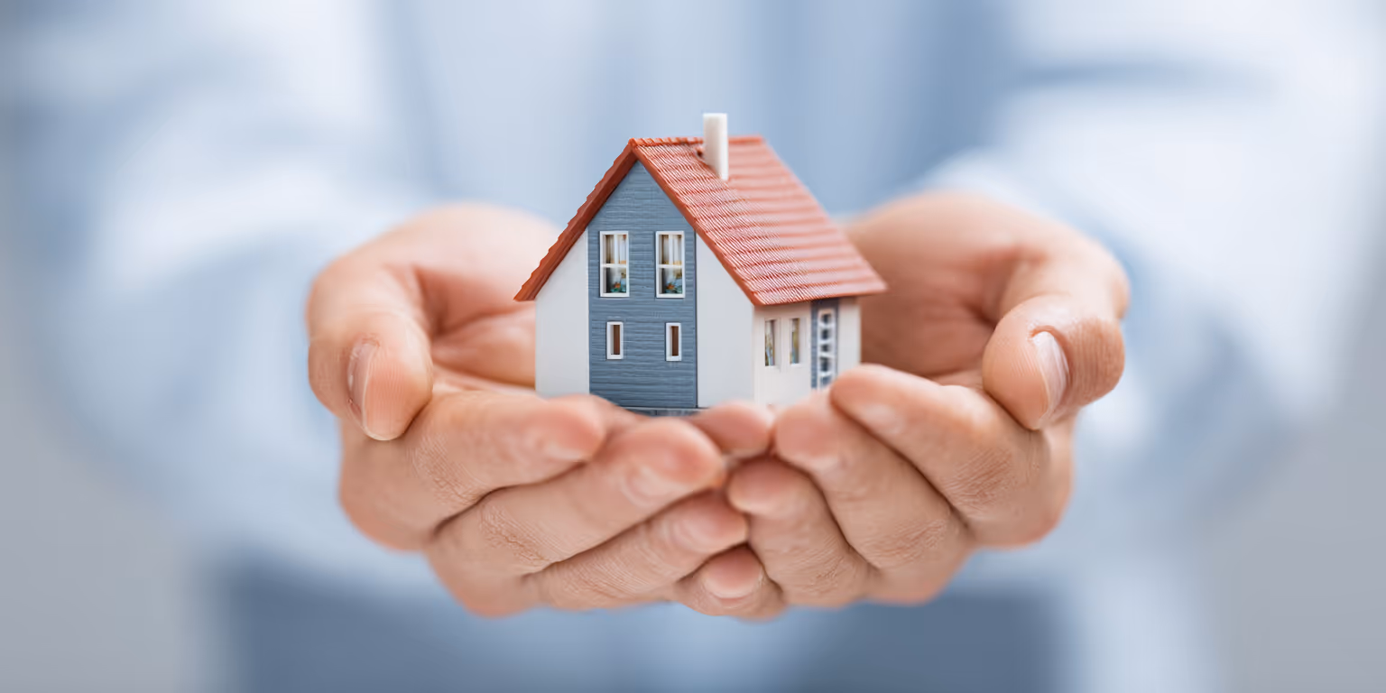 Two hands gently holding a small model house with a red roof and blue and white walls.