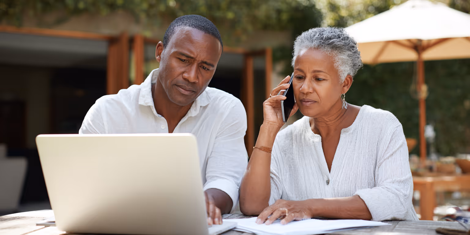 Focused middle-aged man and woman sitting outside at a wooden table, using a laptop and talking on the phone while reviewing papers.