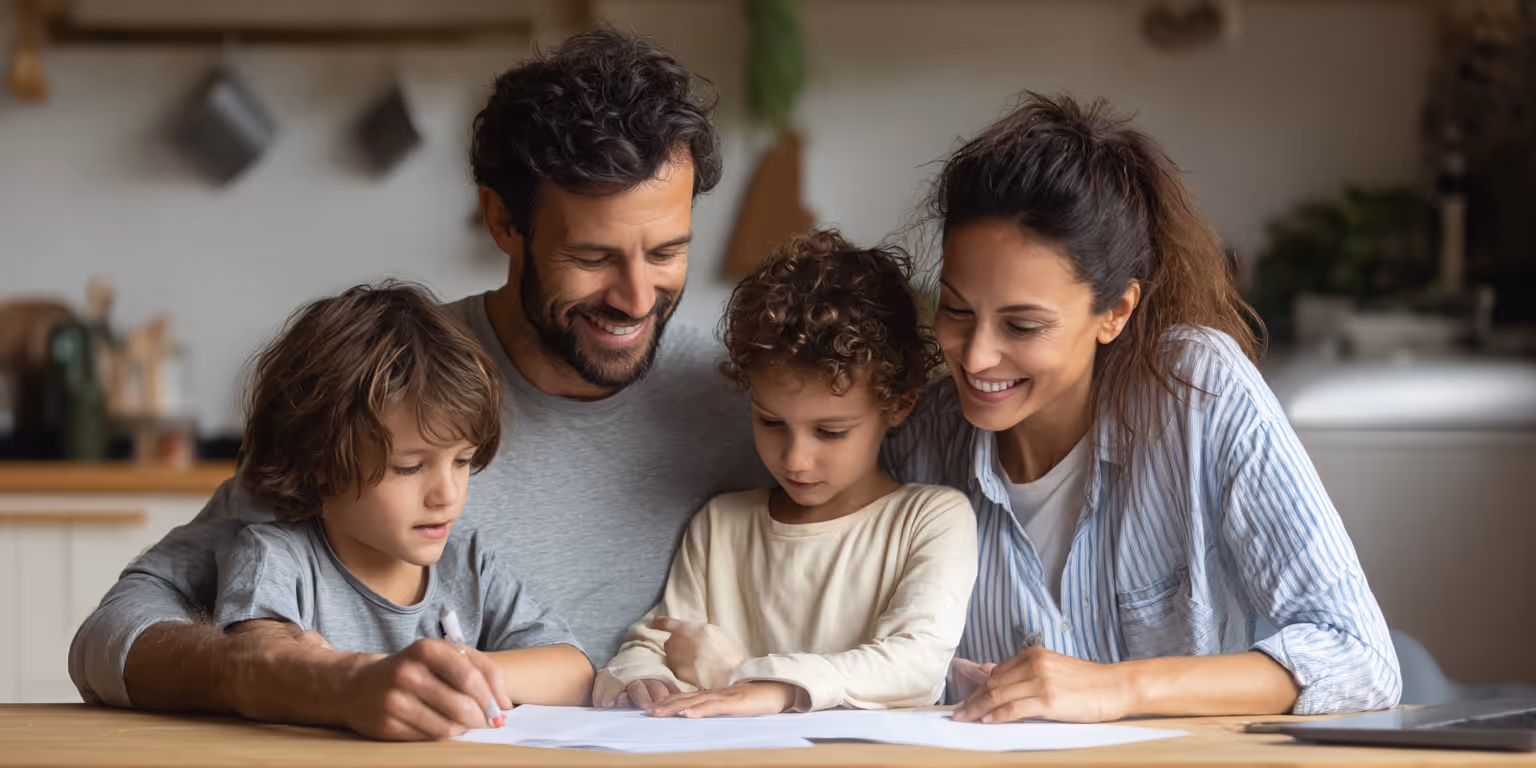 Smiling parents helping their two young children with drawing or homework at a wooden table in a cozy kitchen.