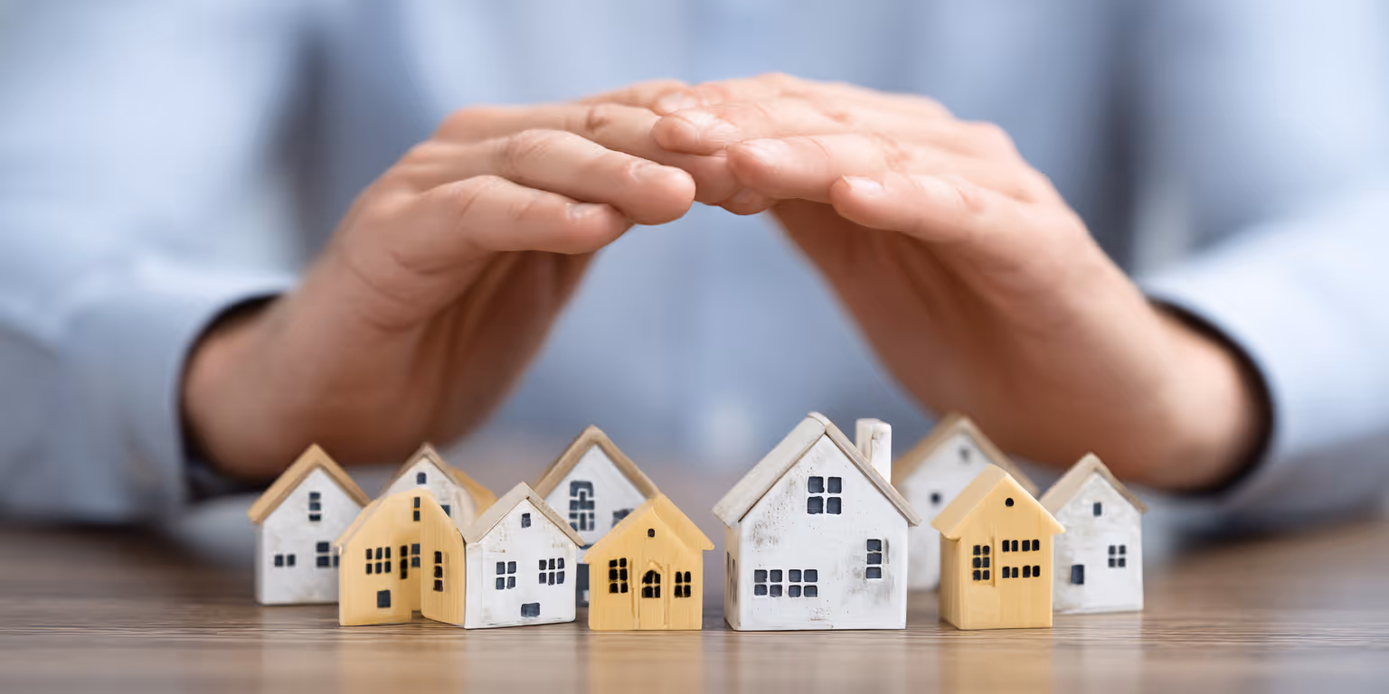 Two hands hovering protectively over a group of small, model houses arranged on a wooden surface.
