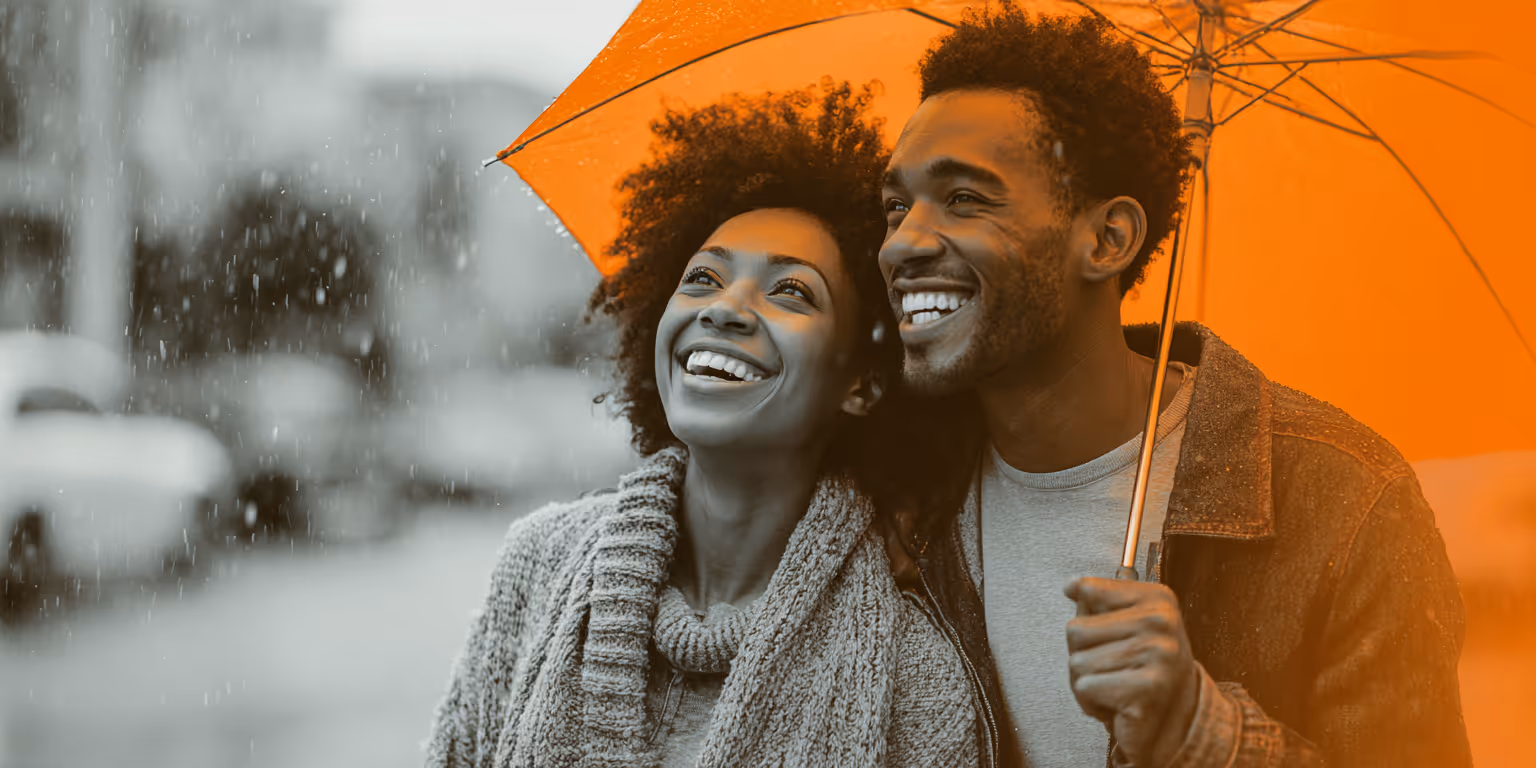Smiling couple standing close under an orange umbrella in the rain.