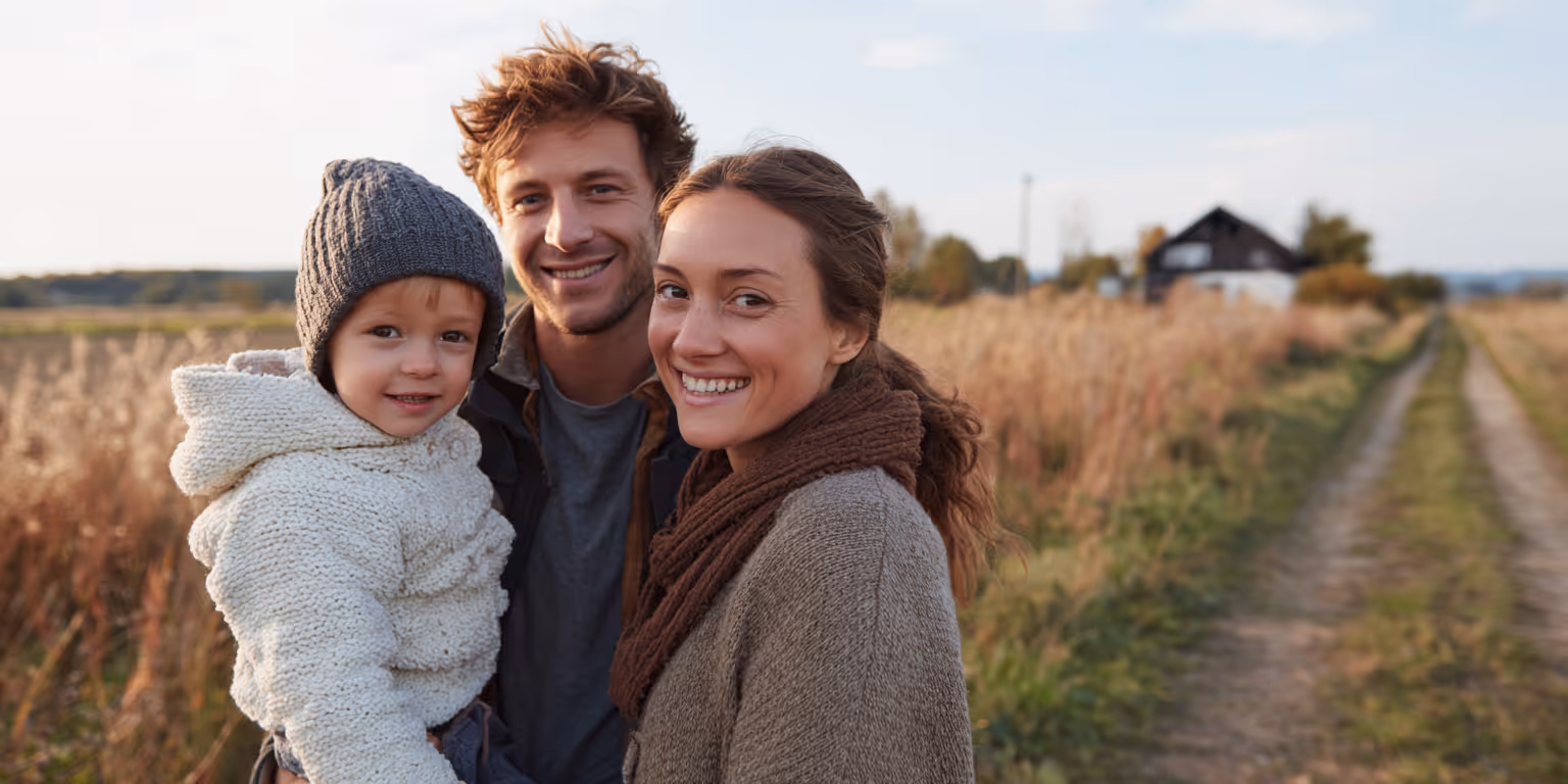 Happy family of three standing close together outdoors with a field and house in the background.