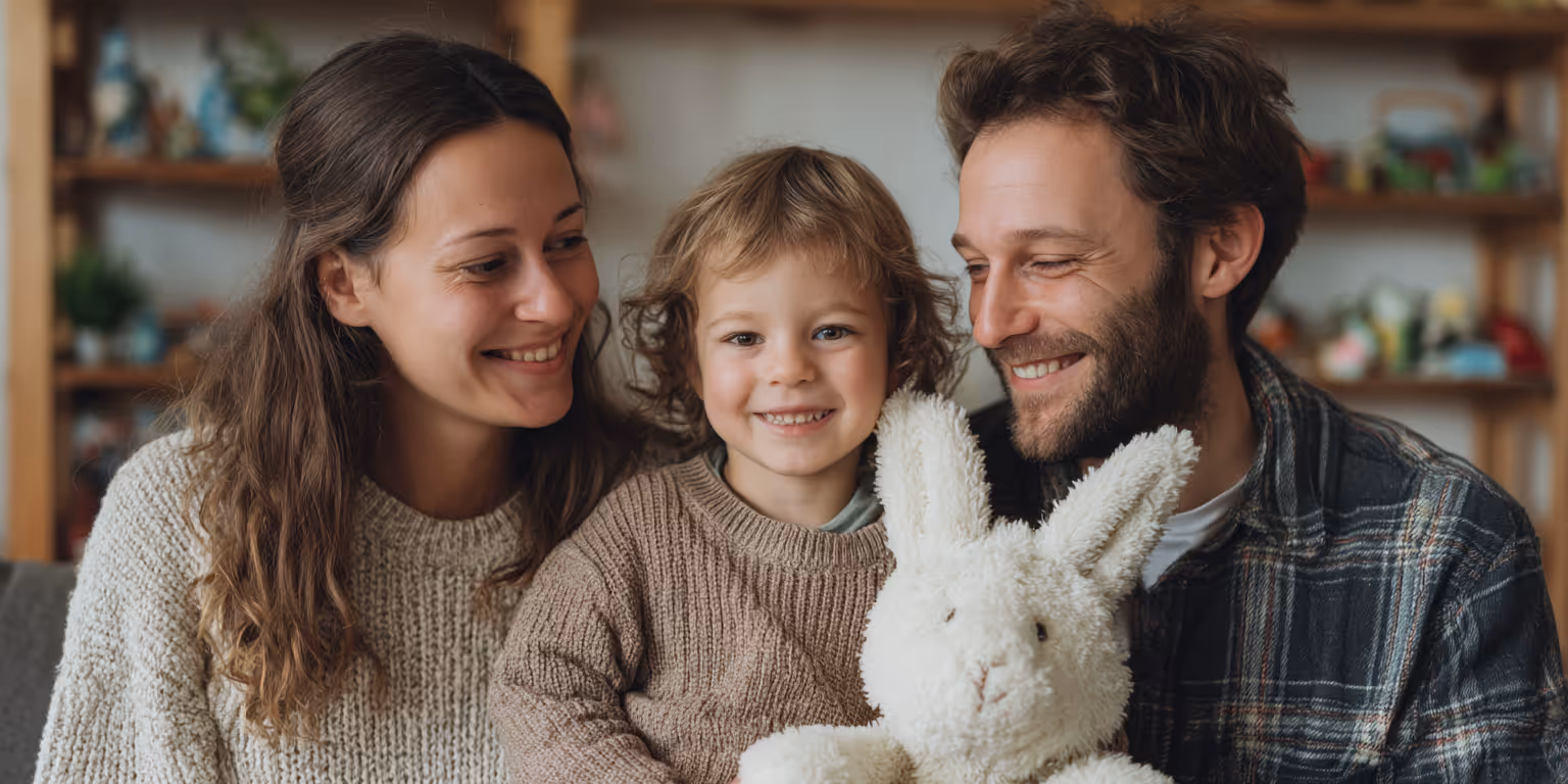 Smiling family of mother, father, and child holding a white stuffed bunny indoors.