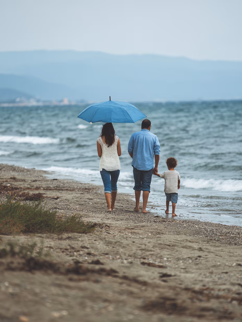 Family of three walking barefoot on a beach near the ocean with the mother holding a blue umbrella.