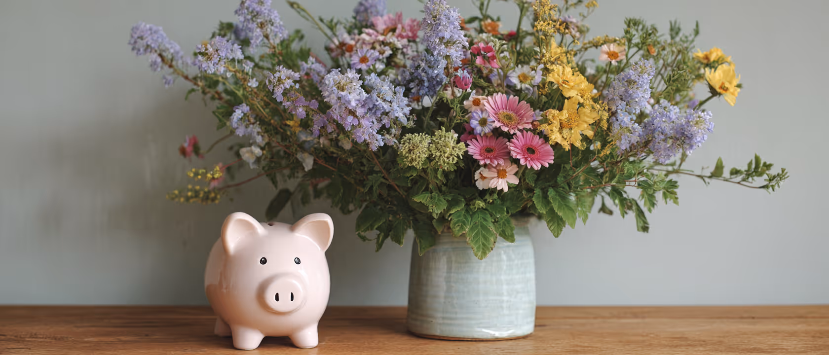Pink ceramic piggy bank next to a large bouquet of mixed fresh flowers in a light blue vase on a wooden surface.