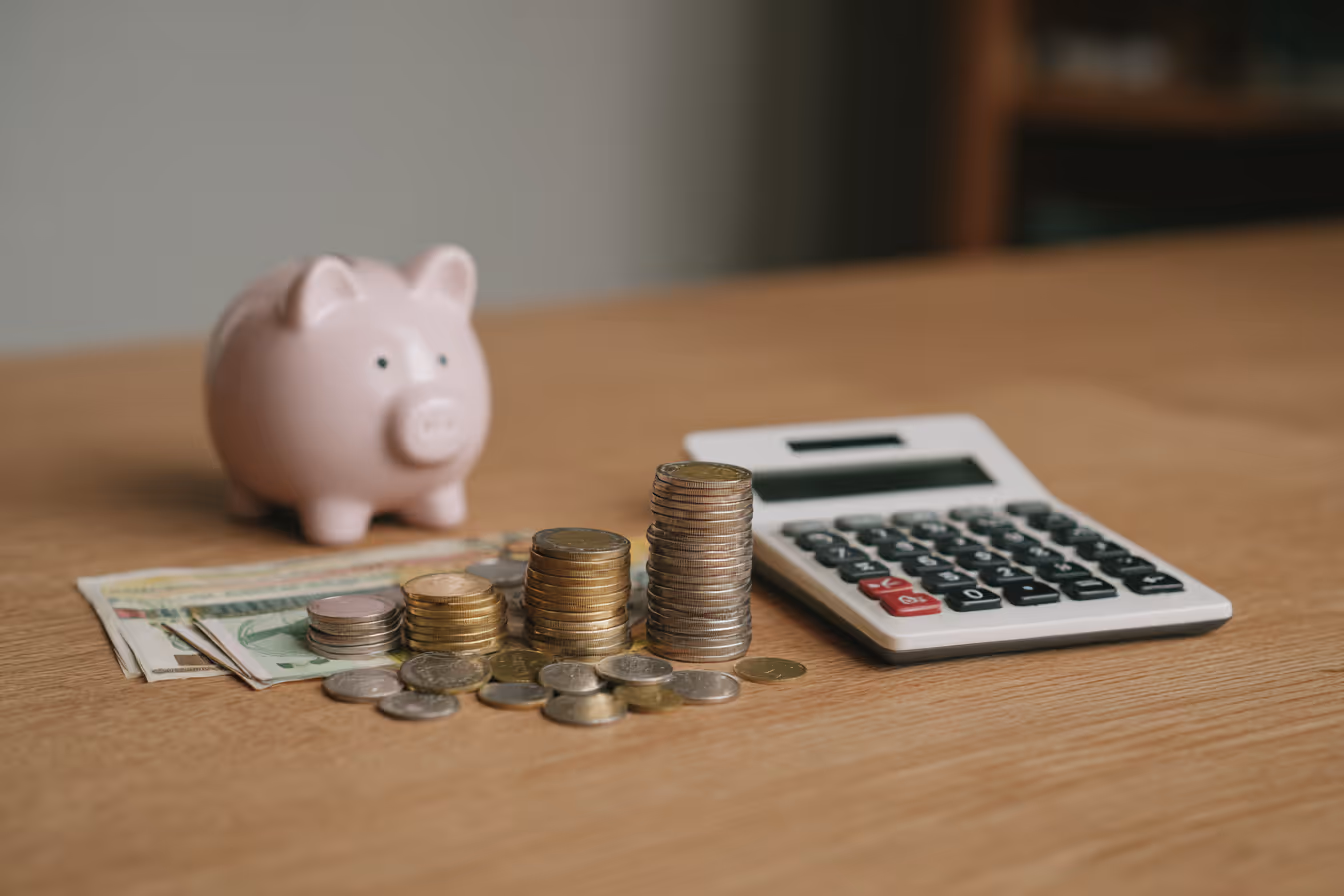 Stacks of coins, a calculator, paper money, and a pink piggy bank on a wooden table.