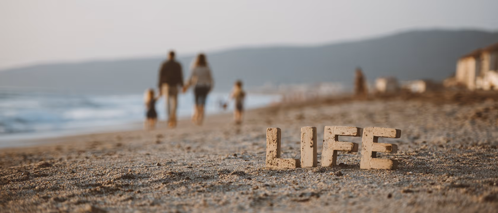 Word LIFE made of wooden letters standing on a sandy beach with a blurred family walking in the background near the water.