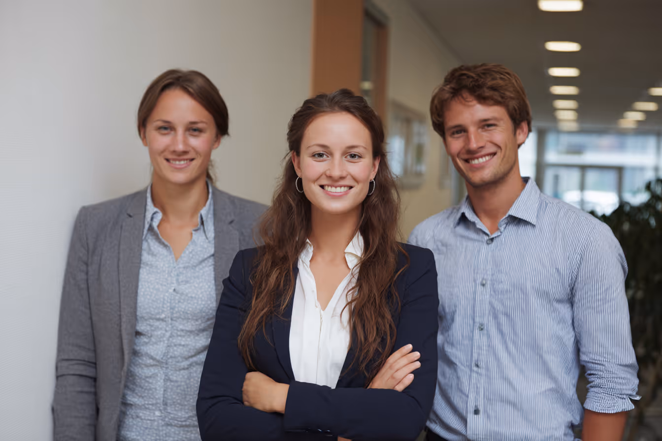 Three young professionals standing in a well-lit office hallway, smiling confidently at the camera.