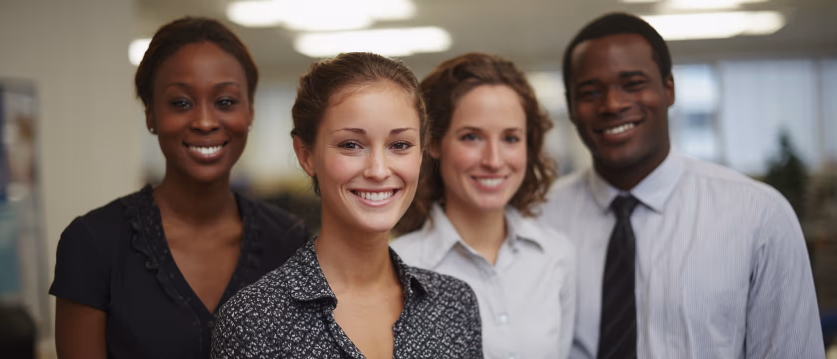 Four diverse professionals smiling in an office setting, two women in front and one woman and one man slightly behind them.
