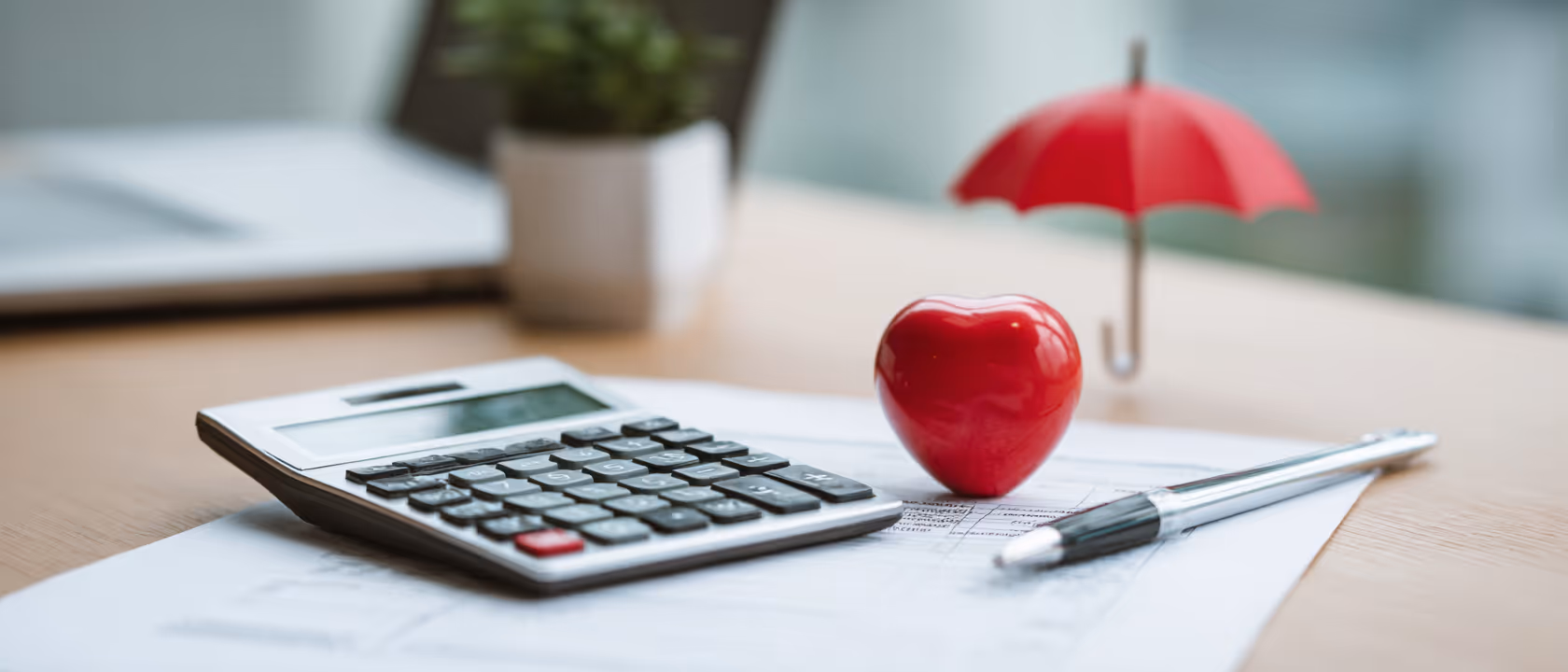 Calculator, pen, and a red heart on paper with a small red umbrella in the background, symbolizing health insurance or financial protection.