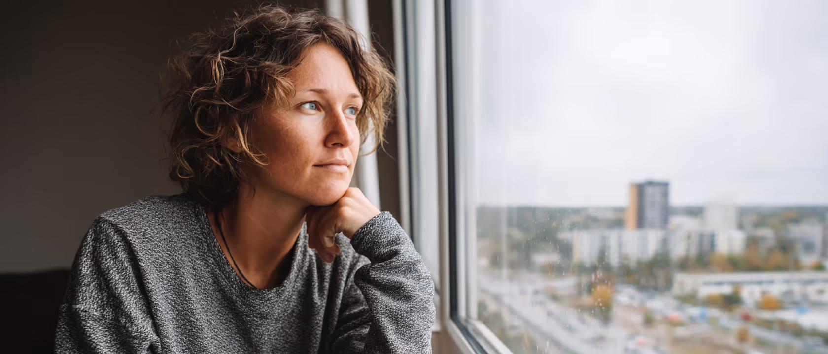 A woman with curly hair in a gray sweater gazing thoughtfully out a window at a cityscape.