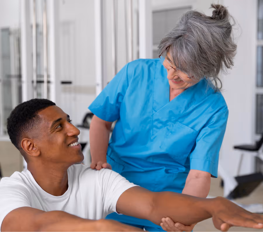 Smiling healthcare professional in blue scrubs assisting a young man stretching his arm in a clinical setting.