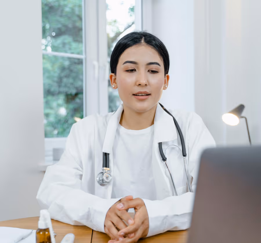 Female doctor wearing a white coat and stethoscope sitting at a desk and speaking in front of a laptop.