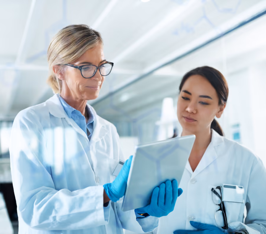 Two female scientists in lab coats looking at a tablet in a bright laboratory.