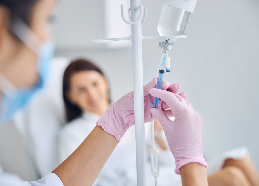 Medical professional wearing pink gloves fills a syringe from an IV drip bag while patient relaxes in the background.