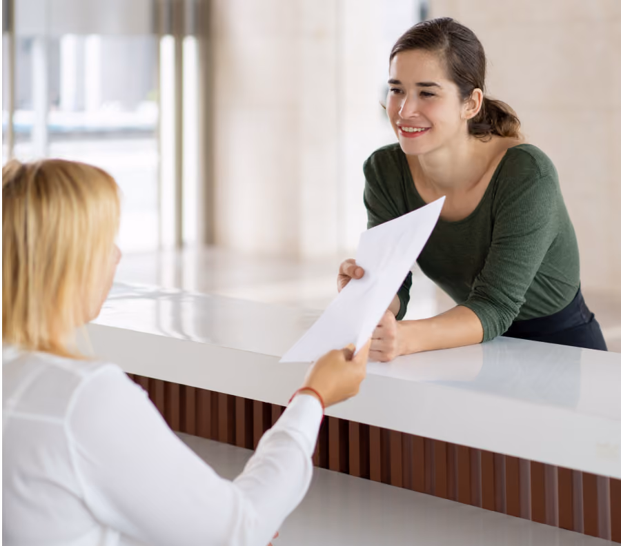Woman in green shirt smiling and receiving a paper from a seated woman at a white reception desk.