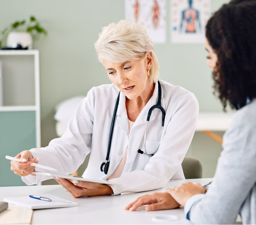 Female doctor in white coat with stethoscope explains something to person across the table.