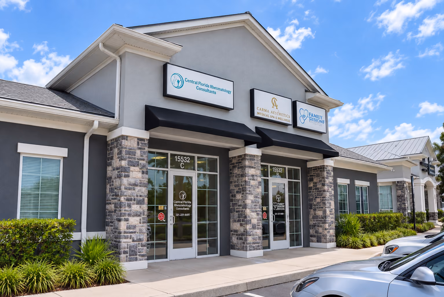 Modern commercial building with stone and gray exterior walls under a blue sky, housing multiple medical businesses including Central Florida Rheumatology Consultants, Carma Aesthetics, and a Family Medicine practice, with parked cars in front.