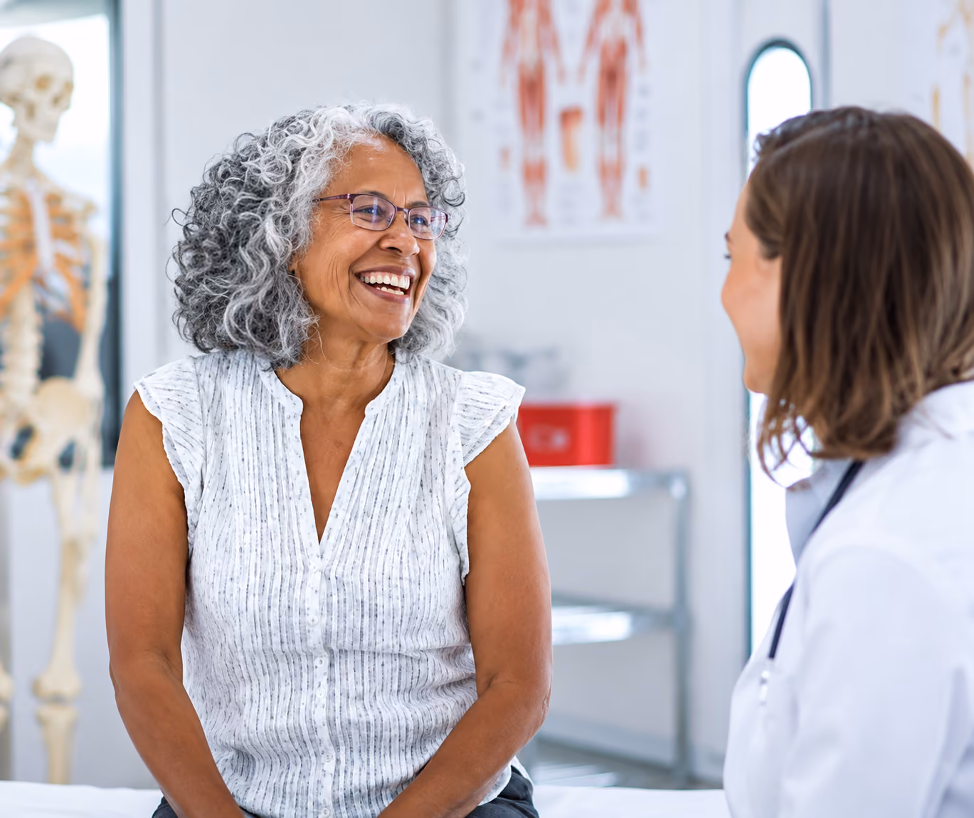 Smiling elderly woman with curly gray hair wearing glasses and a sleeveless blouse talks with a female healthcare professional in a clinical setting with anatomical models in the background.