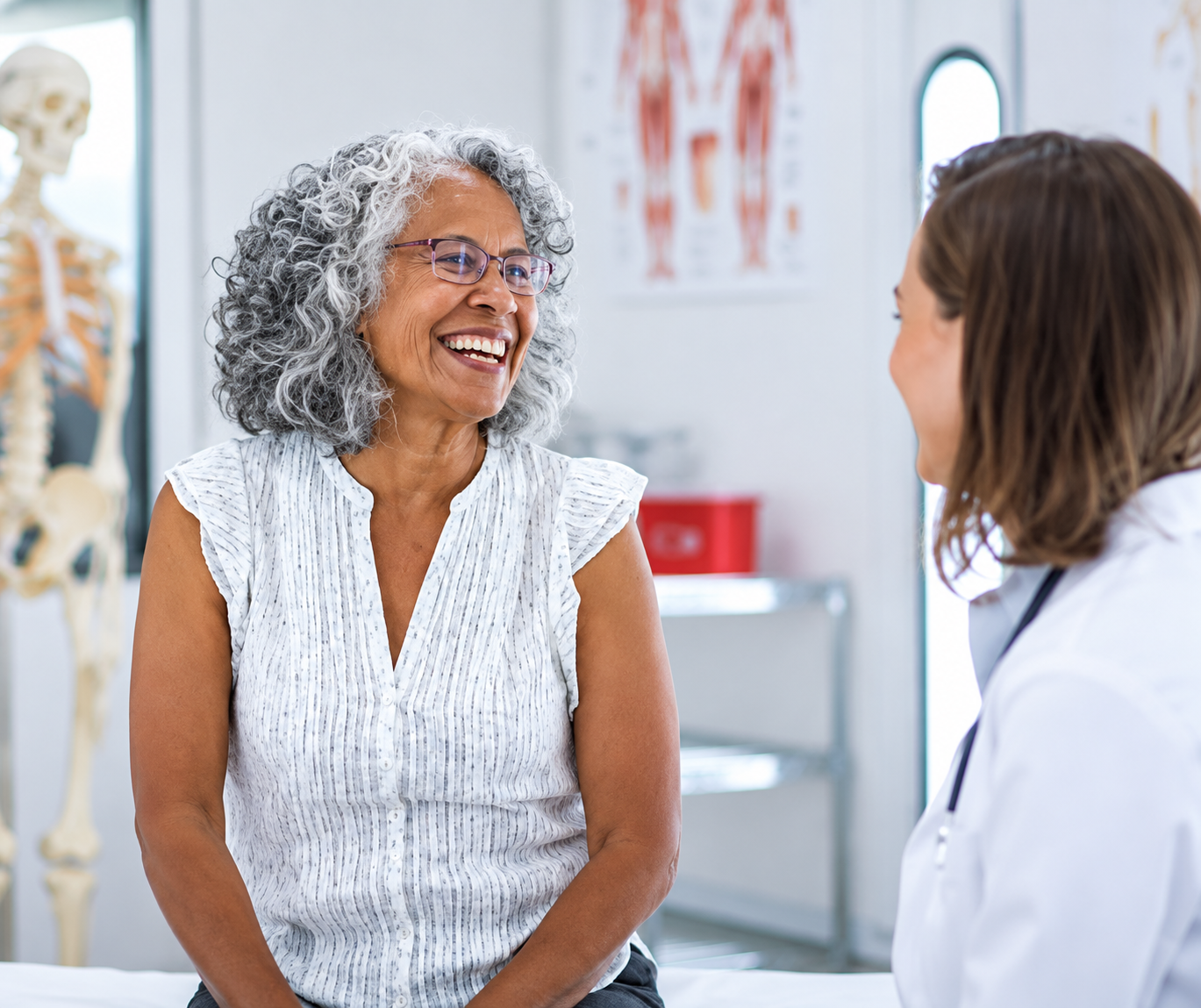 Smiling older woman with curly gray hair and glasses sitting on a medical exam table, talking to a female doctor in a white coat in a bright clinic room with anatomical posters and a skeleton model in the background.