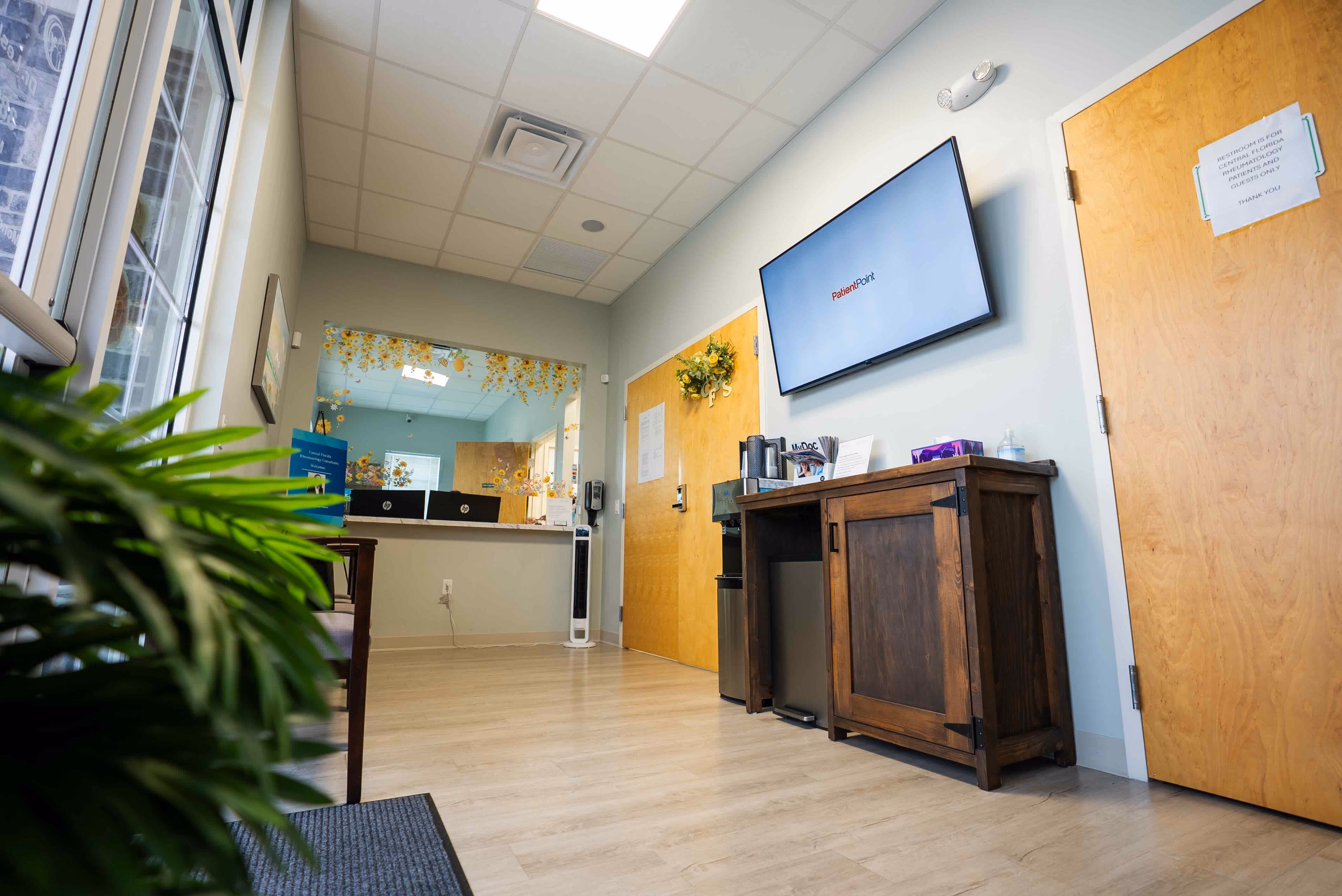 Reception area in a CFRC office with wooden floor, light blue walls, a large window with a green plant in front, a wooden cabinet with informational brochures, a flat screen TV showing 'PatientPoint', and a door with a sign indicating restrooms for patients and guests.