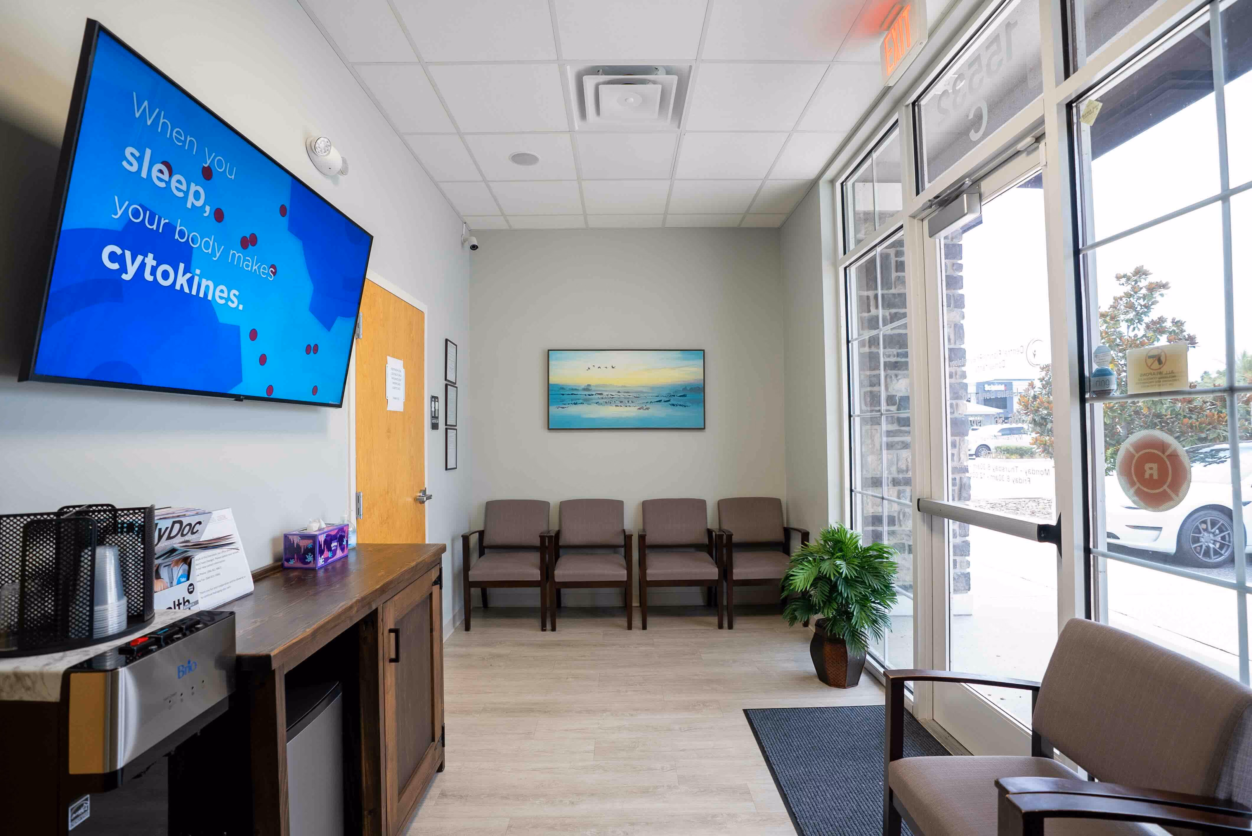 Bright waiting room with five brown chairs along the back wall and one chair near glass entrance doors. A flat screen on the left wall displays text about sleep and cytokines. There is a wooden cabinet with a water cooler, cups, tissues, and pamphlets beneath the screen. A green potted plant is on the floor near the doors, and a landscape painting hangs on the back wall. The room has light wood flooring and a white ceiling with recessed lighting.