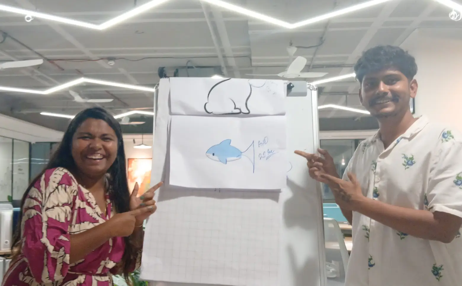 Two people smiling and pointing at a whiteboard with drawings of a blue fish and a white animal outline in an office setting.