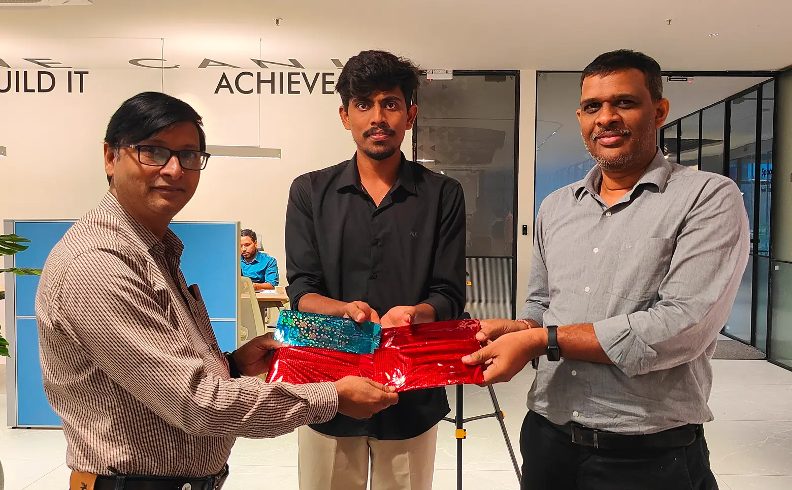 Three men standing in an office holding shiny wrapped gift packages, with two men on either side holding the gifts and the man in the center supporting them.