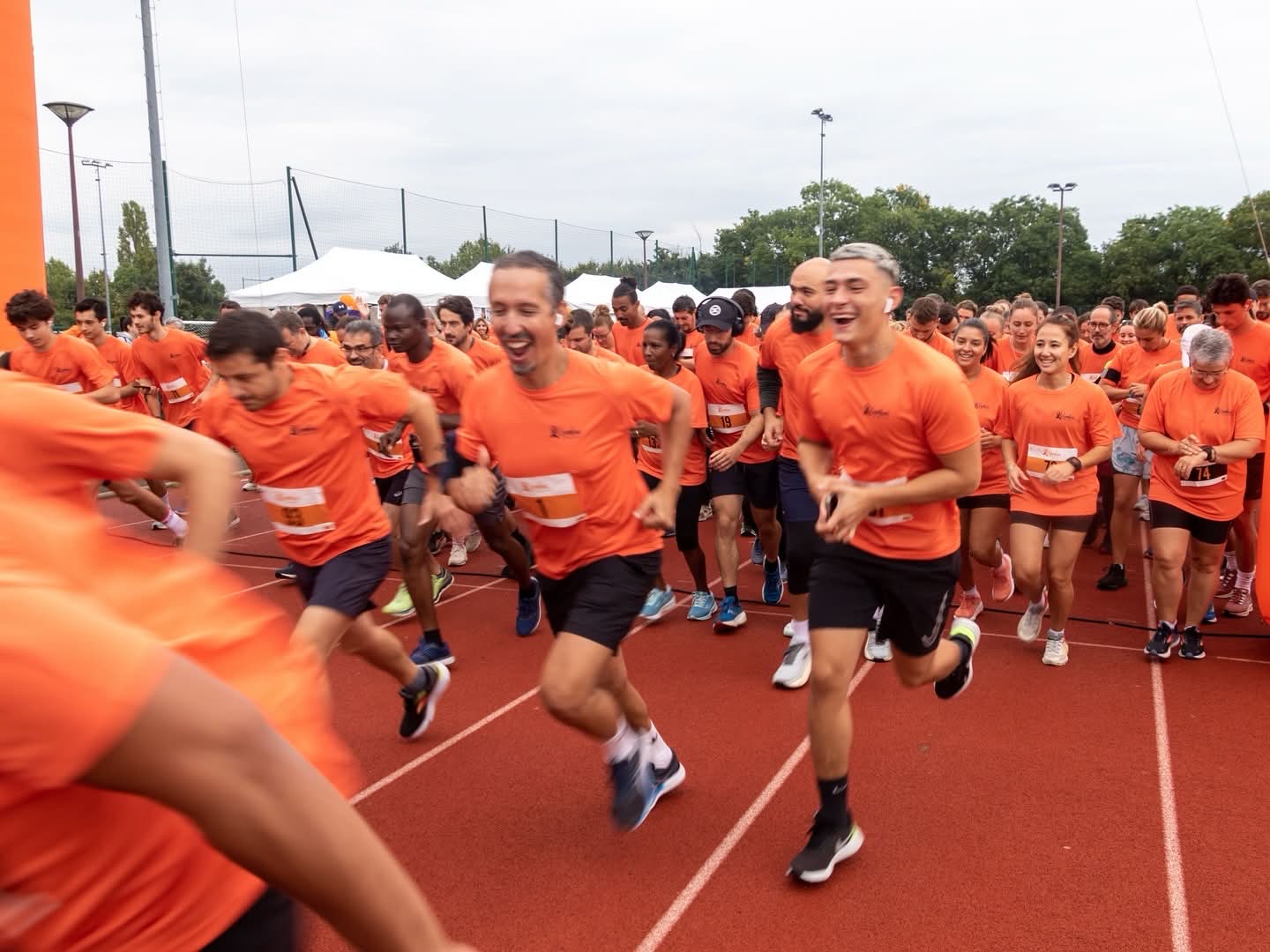 Groupe de coureurs souriants portant des t-shirts orange démarrant une course sur une piste extérieure.