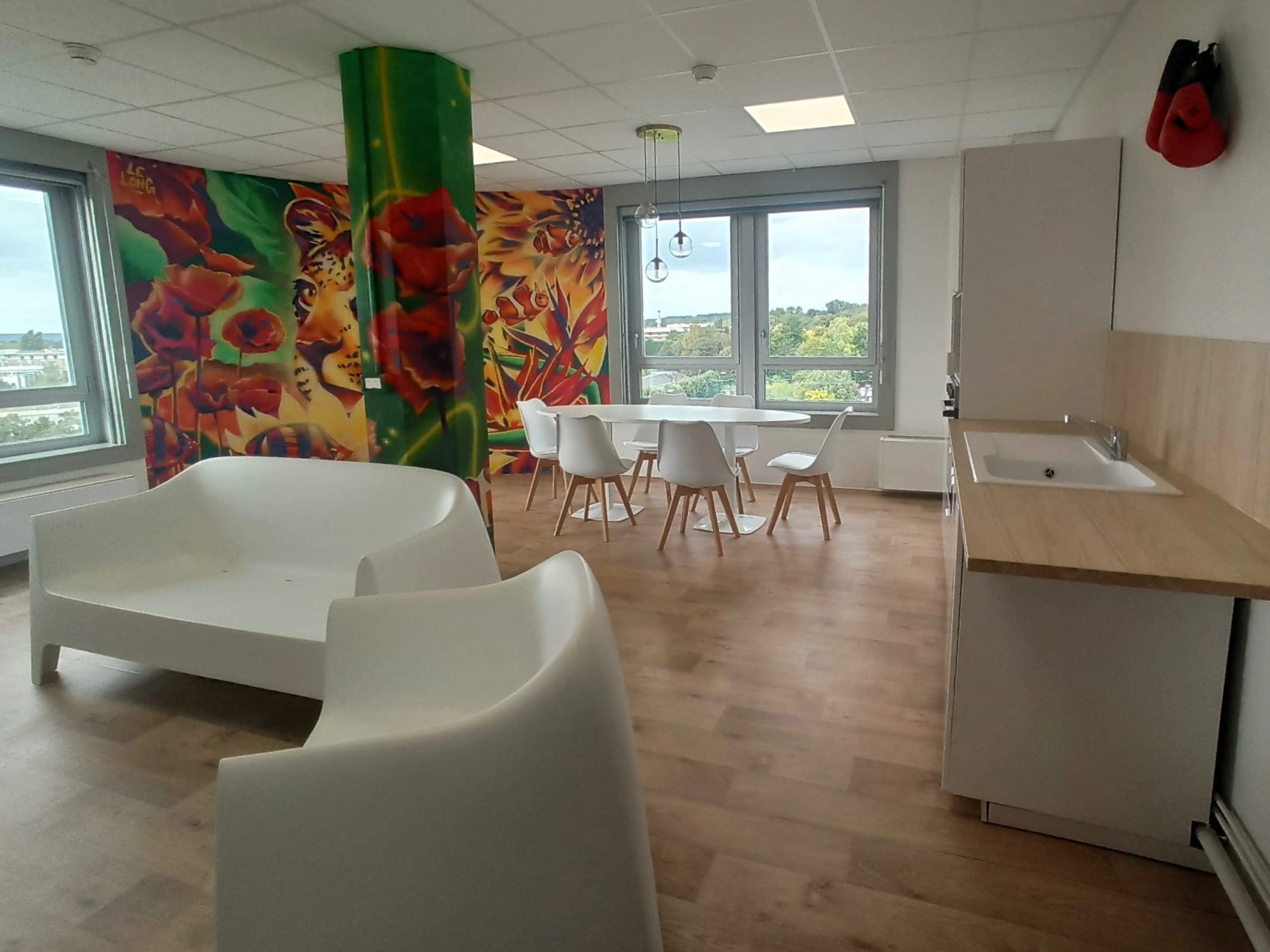 Salle de repos avec canapés blancs, table ronde avec chaises blanches, cuisine équipée, mur peint avec tigre et fleurs, et paire de gants de boxe rouges suspendue.