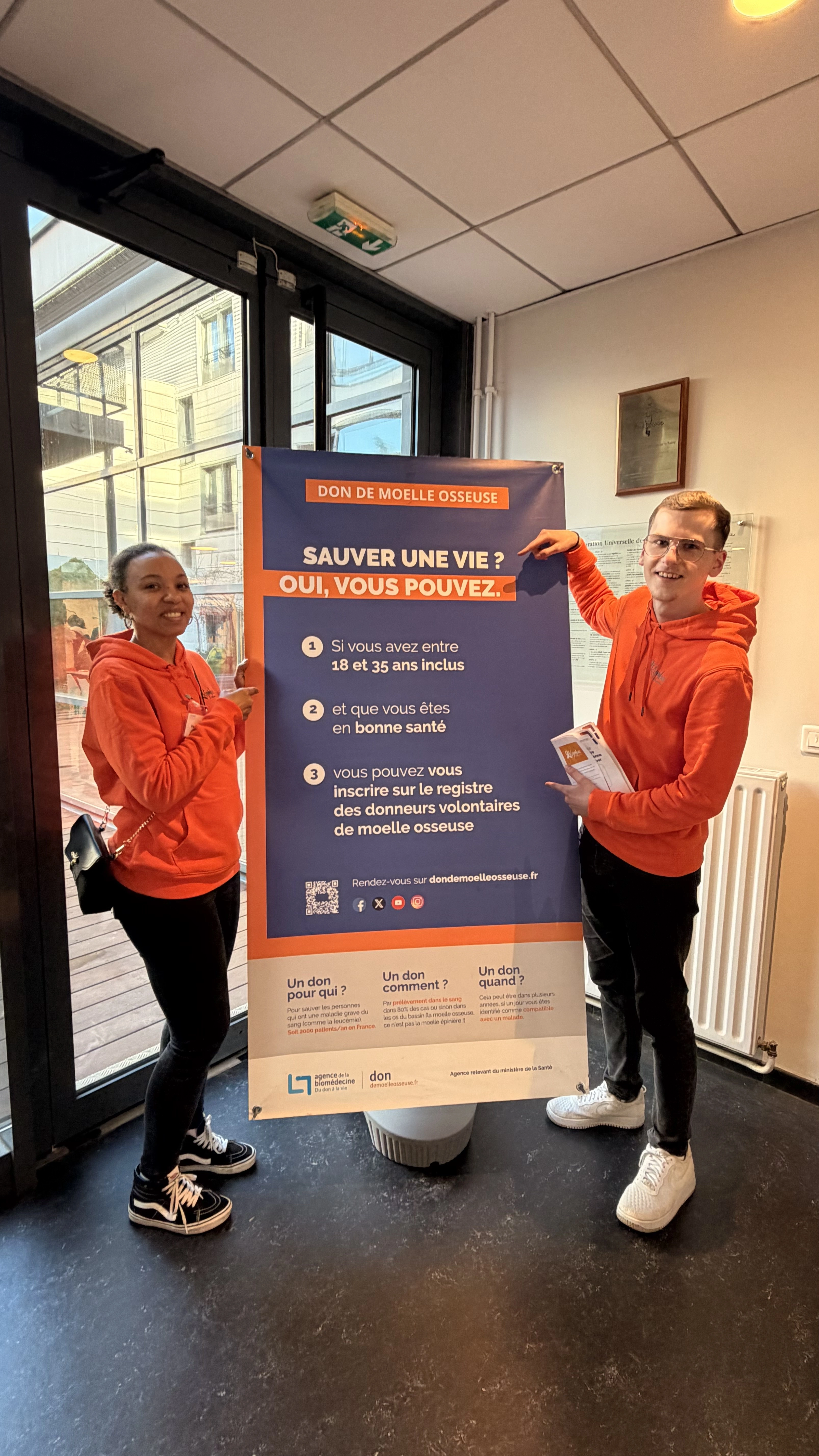 Deux jeunes adultes en sweat à capuche orange tenant une affiche bleue et orange sur le don de moelle osseuse avec des informations et un QR code.