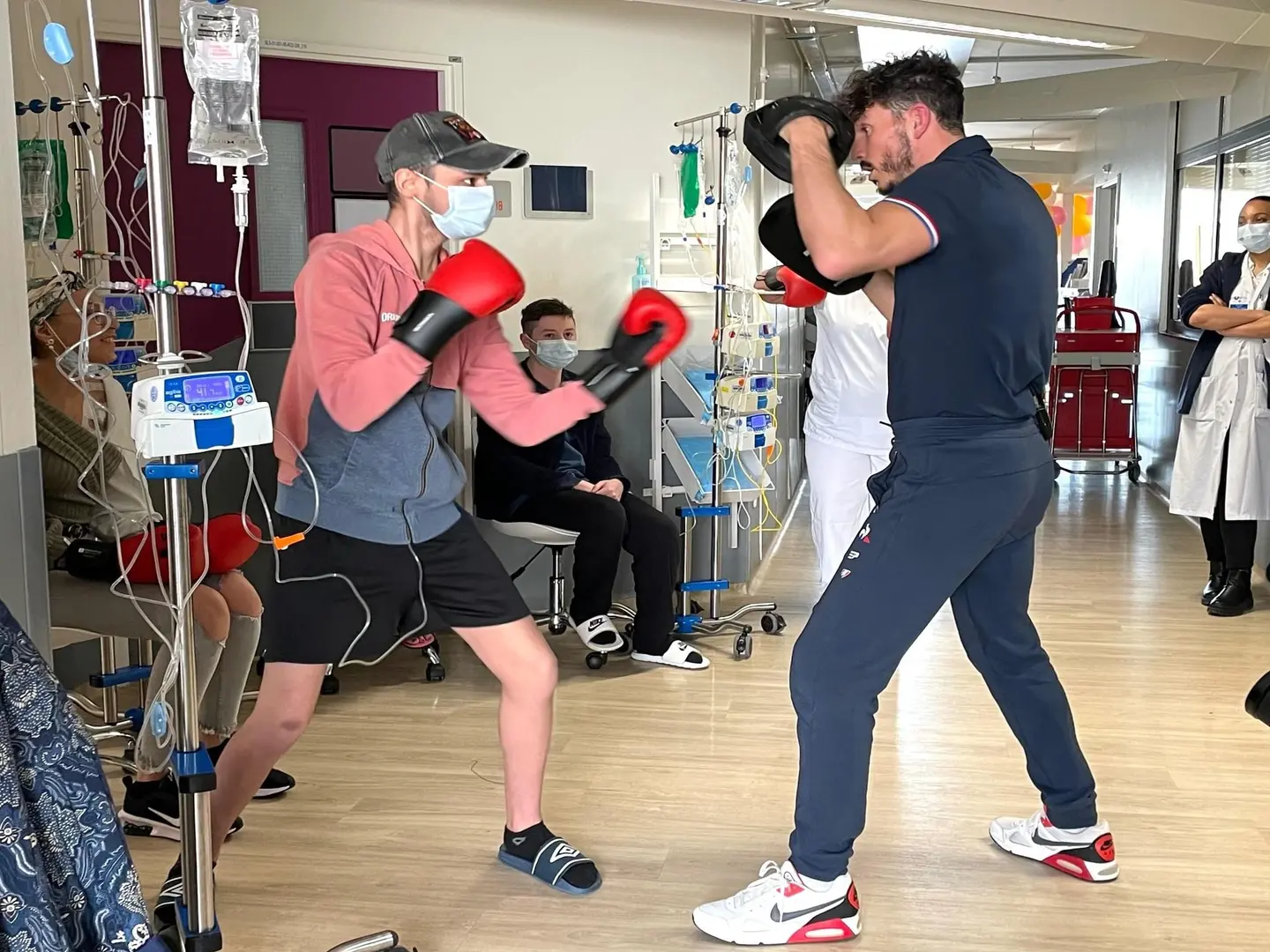 Un patient masqué en gants de boxe rouges s'entraîne en boxe avec un entraîneur dans un couloir d'hôpital, entouré de personnel médical et d'autres patients.