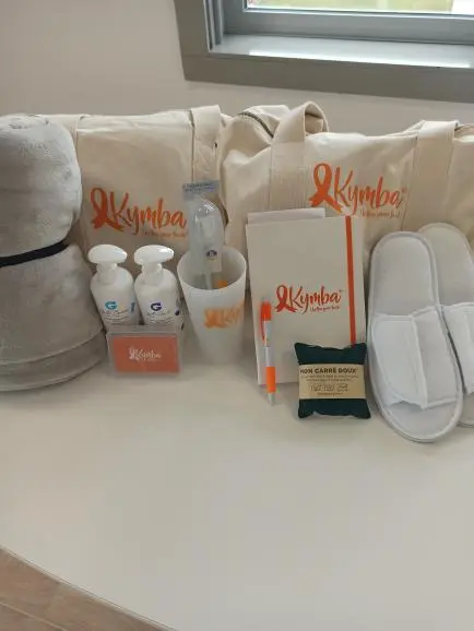 Ensemble d’articles de confort comprenant serviette grise roulée, sacs à linge crème avec logo orange Kymba, flacons de lotion, carnet, stylo, savon naturel, et paires de chaussons blancs.