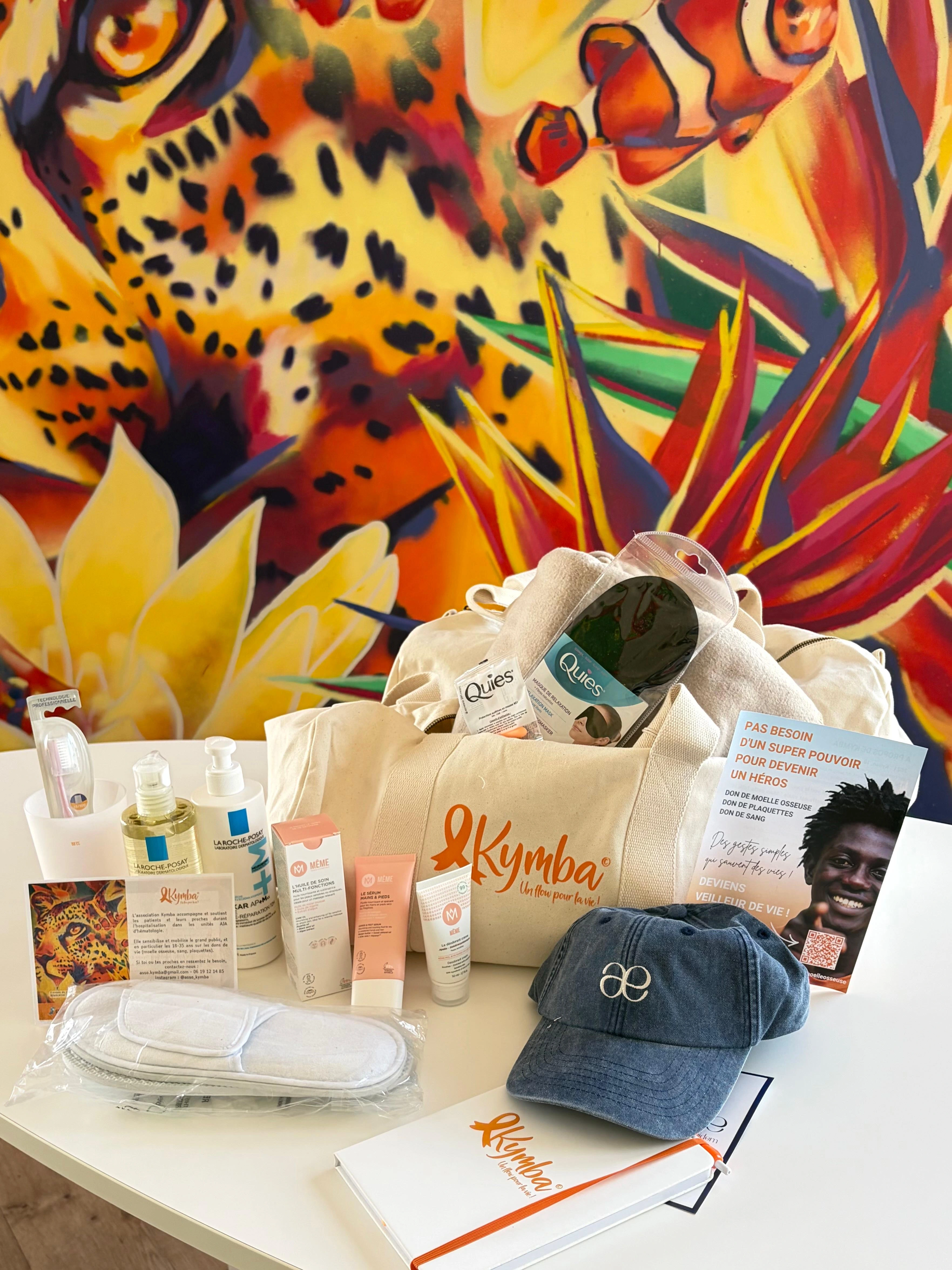 Ensemble d’articles de confort comprenant serviette grise roulée, sacs à linge crème avec logo orange Kymba, flacons de lotion, carnet, stylo, savon naturel, et paires de chaussons blancs.