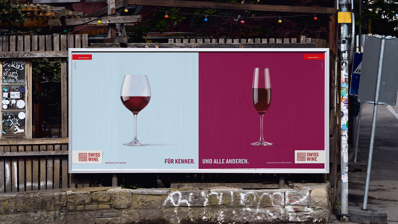 Swiss Wine Grossplakat an einer Strasse: links ein bauchiges Rotweinglas auf Hellblau, rechts eine schlanke Flöte auf Bordeaux – «Für Kenner. Und alle anderen.»