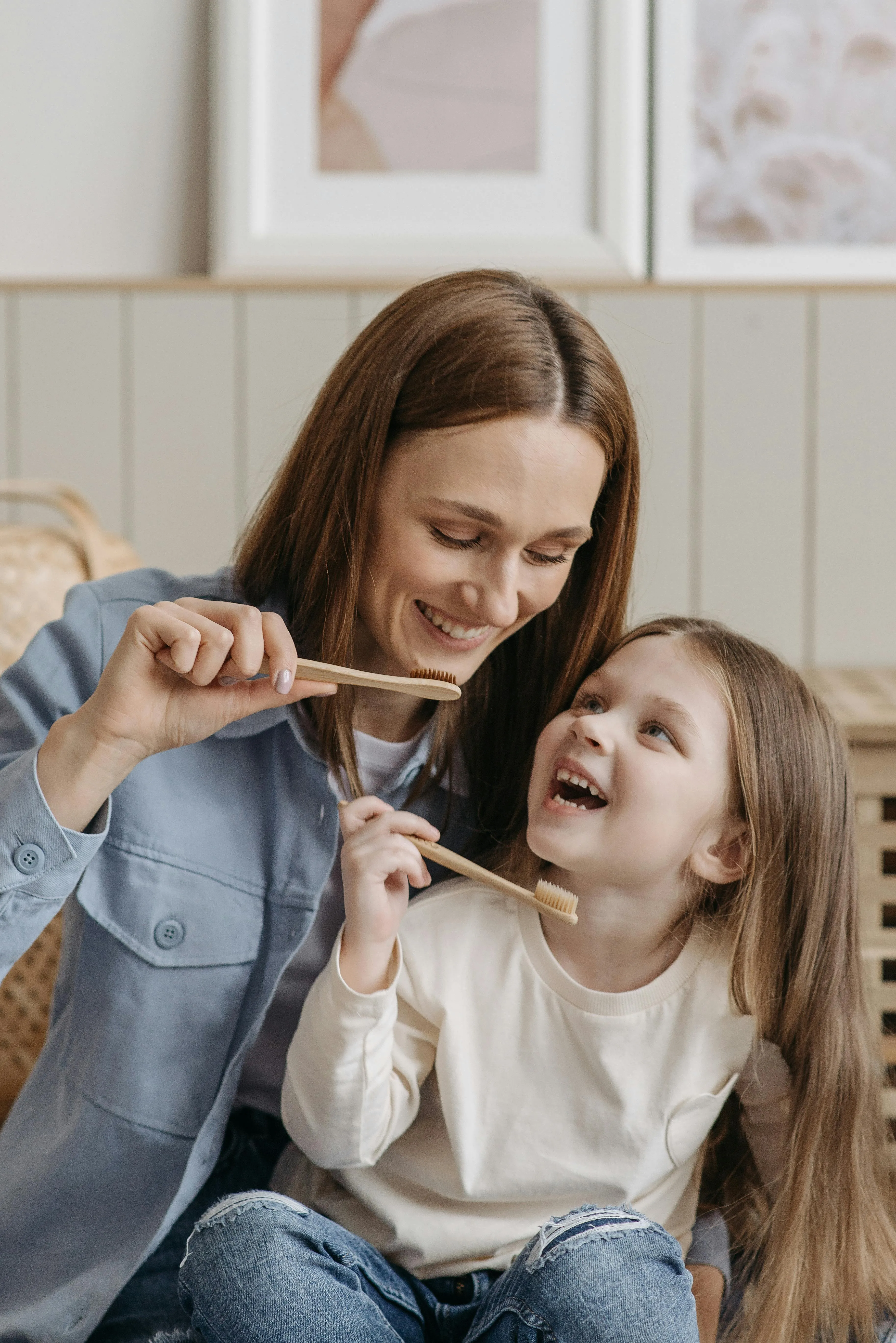 Mother smiling while helping her young daughter brush her teeth with a wooden toothbrush, illustrating at-home oral health guidance for parents
