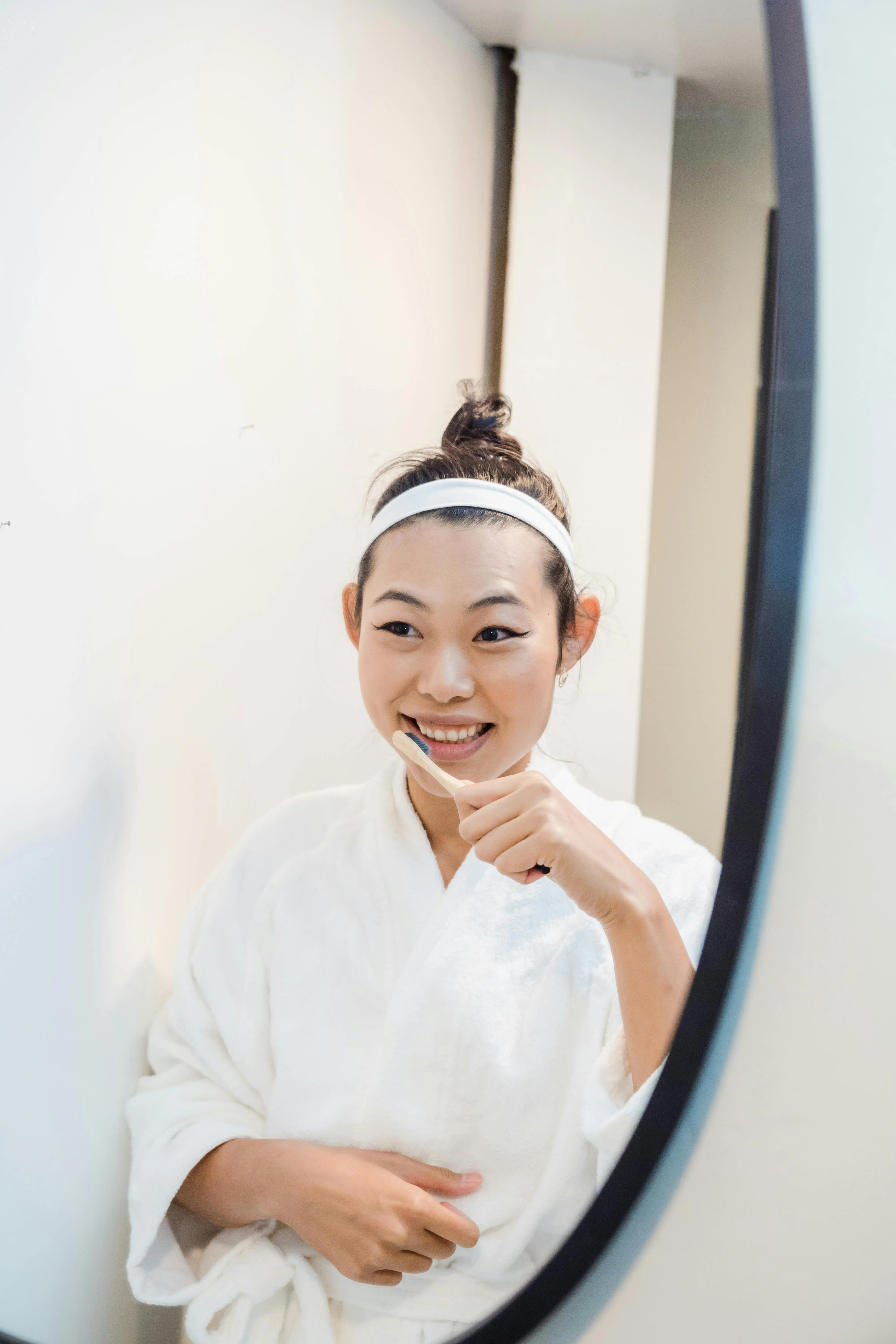 Smiling woman with a white headband brushing her teeth in front of a mirror, representing the importance of proper aftercare following a tooth extraction