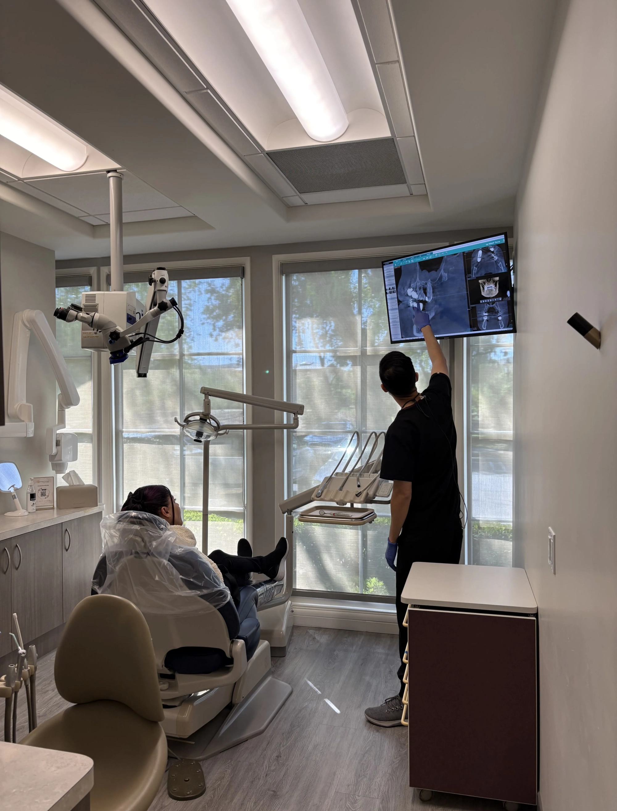 Dr. Kim pointing to a patient's dental X-rays displayed on an overhead monitor in the Ortega Cottage Dentistry exam room, with dental microscope visible