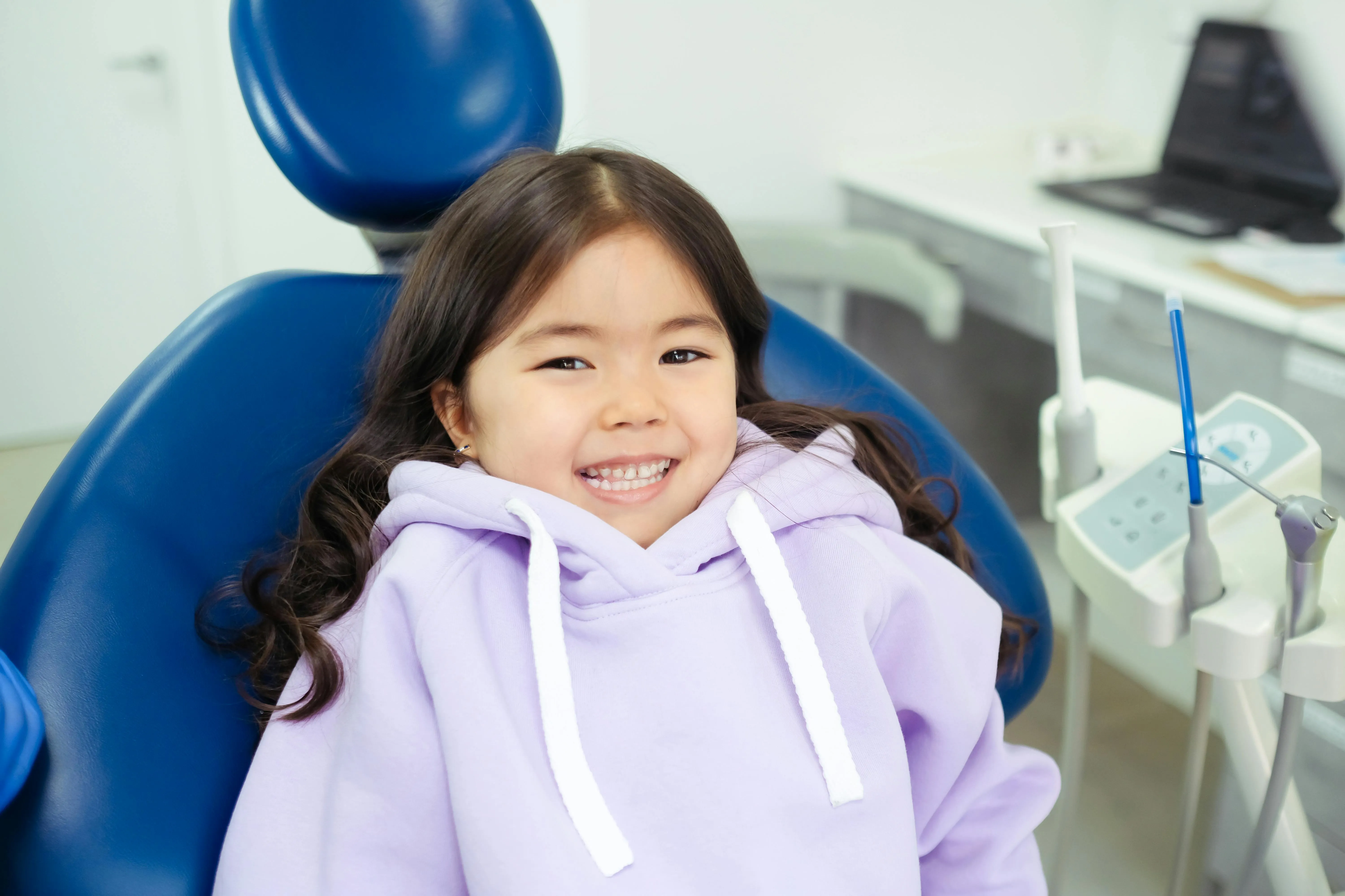 Smiling young girl in a purple hoodie sitting in a blue dental chair at a dentist office.